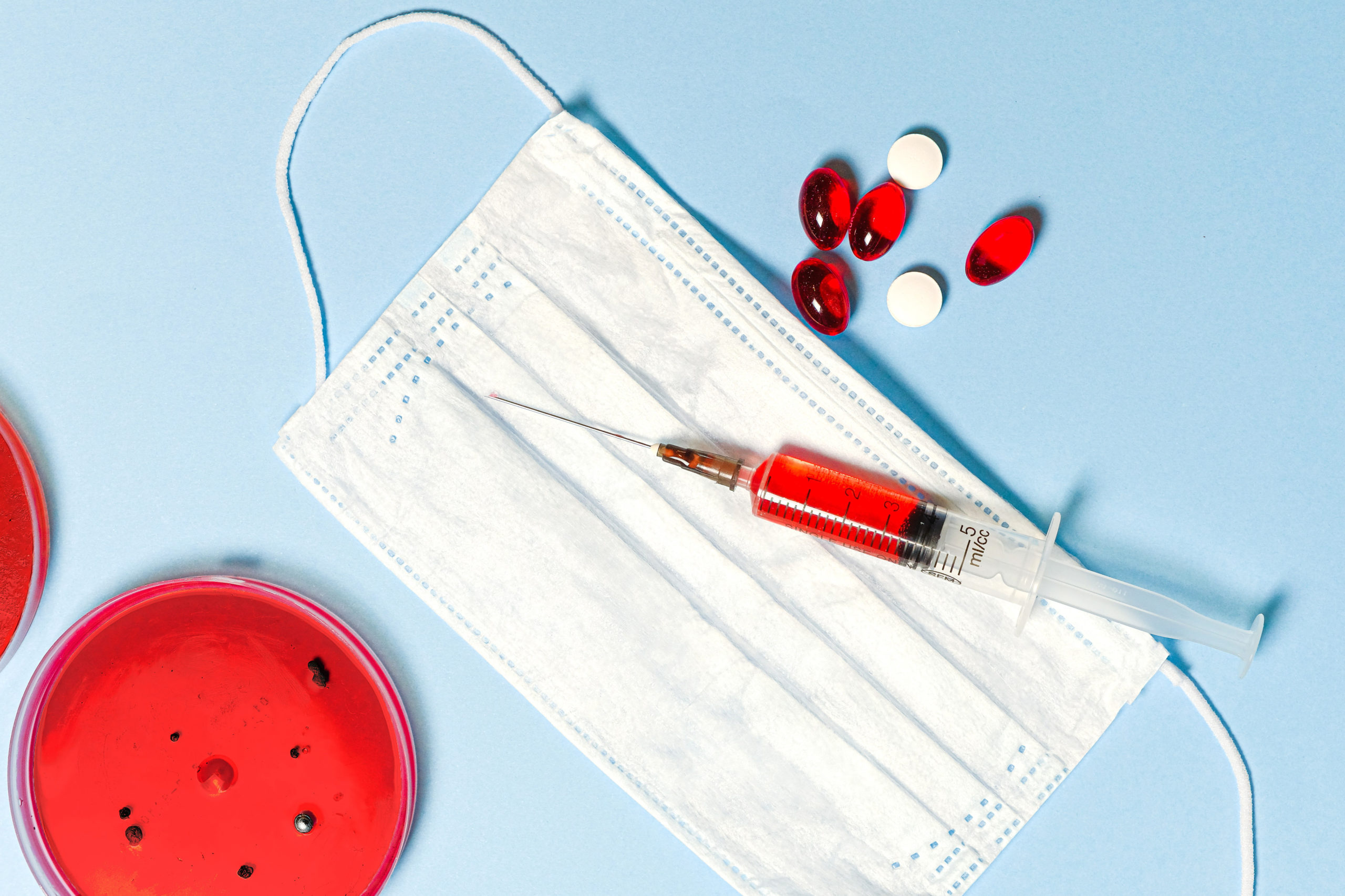 Zdjęcie przedstawiające strzykawkę z czerwonym płynem, szalkę petriego z wyhodowanymi bakteriami, maseczkę jednorazową oraz medykamenty - badania laboratoryjne w Krzyżu Wielkopolskim
